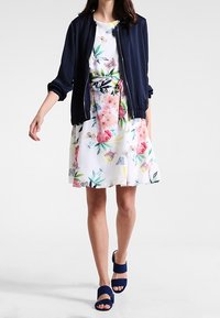 Robe florale avec des motifs colorés sur fond blanc, associée à une veste bomber bleu marine et des sandales ouvertes bleues.