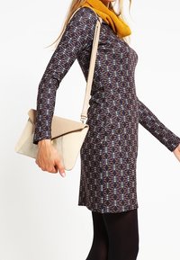 Abito a maniche lunghe di colore blu scuro con un motivo geometrico, abbinato a una borsa a tracolla beige. La sciarpa è di colore senape, creando un contrasto.