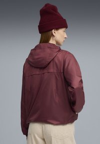 Bordeauxfarbene leichte Jacke mit Kapuze, glatter Textur, elastischen Bündchen und einer Nahtdetail auf der Rückseite, kombiniert mit einer gestrickten bordeauxfarbenen Mütze.