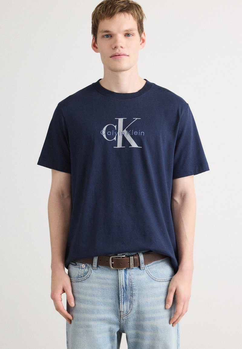 Námornícky modré bavlnené tričko s strieborným logom Calvin Klein. Krátke rukávy, dizajn s okrúhlym výstrihom, skombinované s svetlými denimovými džínsy a hnedým opaskom.