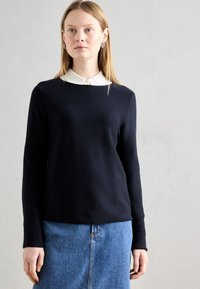 Temno modra jopica z okroglo ovratnico in dolgimi rokavi, prekrita z belim srajčnim ovratnikom. Mehak na dotik, preprost dizajn, minimalno okrasitev.