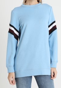 Felpa a maniche lunghe blu chiaro con strisce nere, bianche e bordeaux sulle maniche superiori, indossata con jeans blu.