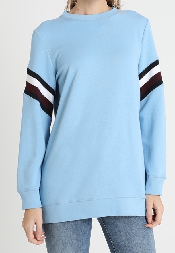 Felpa a maniche lunghe blu chiaro con strisce nere, bianche e bordeaux sulle maniche superiori, indossata con jeans blu.
