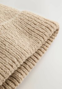 Beige Strickmütze mit einem strukturierten, gerippten Muster. Das Wollmaterial hat ein weiches, fuzzy Aussehen und zeigt detaillierte Nähte.