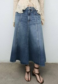 Denim rok met een uitlopende, A-lijn vorm, lichtblauwe kleur en een ruwe zoom. Gecombineerd met bruine sandalen met bandjes. Een textuur van een kanten top is zichtbaar erboven.