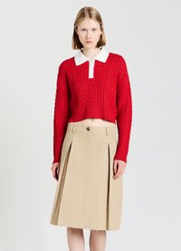 Pull court en maille torsadée rouge avec col blanc, associé à une jupe midi plissée beige avec une fermeture à boutons sur le côté.
