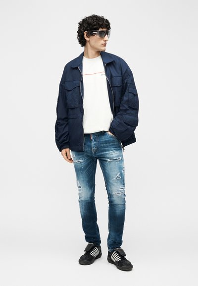 Νέος άνδρας που φοράει υπερμεγέθη σκούρο μπλε μπουφάν, λευκό μπλουζάκι, ξεφτισμένο μπλε τζιν, μαύρα αθλητικά παπούτσια και μεγάλα σκούρα γυαλιά ηλίου, κοιτάζοντας προς τα πλάγια.