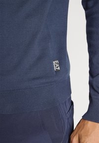 Sötétkék kötött pulóver, gumírozott mandzsettákkal és aljjal, oldalán egy kis fehér címkével, amin az "EA7" felirat található. Texturált anyag és illeszkedő dizájn.
