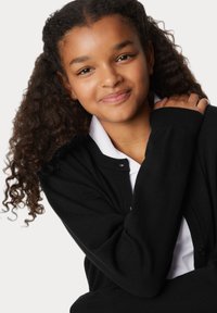 Czarny kardigan z miękkiego włókna, z prążkowanymi mankietami i kołnierzem, noszony na białej koszuli. Tekstura wydaje się gładka i przytulna.