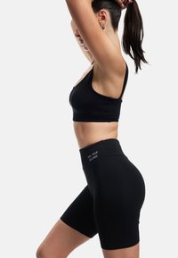 Nainen sitomassa pitkää hiustaan, yllä musta urheiluliivi ja mustat korkeavyötäröiset pyöräilyshortsit, joissa on "TUNNE TUNTEESI" -teksti vyötäröllä.