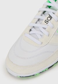 Baskets blanches avec une tige en daim et cuir, lacets blancs, accents verts et semelle à motifs. Le design comprend des détails de couture.