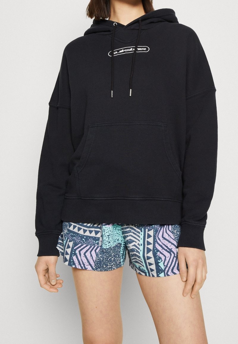 Personne portant un sweat à capuche noir avec poche avant et un short graphique aux motifs géométriques bleus, roses et verts sarcelle, debout devant un fond uni.
