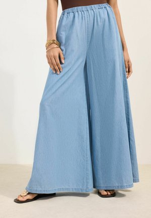 Naine, kellel on seljas helesinised laiade säärtega teksapüksid, pruun top, kuldsed käevõrud ja mustad sandaalid dekoratiivsete kivikestega, seisab beežil põrandal.