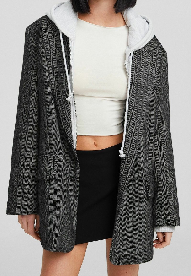 Blazer oversized a spina di pesce grigia con una felpa con cappuccio grigia a contrasto sotto. Visibile un top corto e una gonna nera.