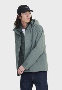 Veste imperméable vert clair avec capuche, dotée de boutons-pression, texture lisse et design décontracté. Portée par-dessus un sweat-shirt gris. Casquette noire.