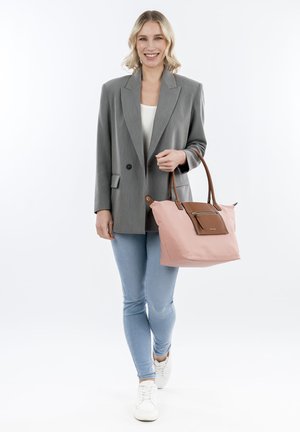 Růžová taška přes rameno s hnědými detaily a zipem. Měkký materiál, prostorný design a dvojité ramenní popruhy. Model má na sobě šedý blazer a džíny.