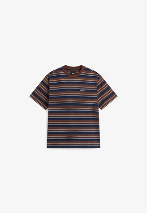 Junger Mann mit langen Haaren, der ein braun-navy gestreiftes Vans-T-Shirt und beige Hosen trägt, steht vor einem schlichten Hintergrund.