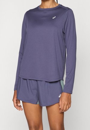 Person trägt ein langärmeliges violettes Sportshirt und passende Shorts mit weißen ASICS-Logos auf Brust und linkem Bein.