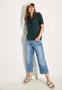 Paita tummanvihreä lyhythihainen, vaaleansiniset leveälahkeiset farkut ja hopeiset sandaalit soljella. Mallilla on päällään beige lippalakki. Yksinkertainen tausta.