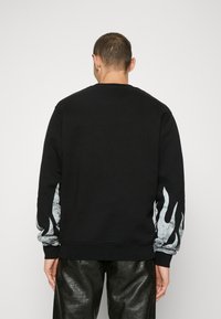 Svart sweatshirt med vit flamgrafik på ärmarna, med ribbade muddar och nederkant, tillverkad av mjukt, texturerat tyg.