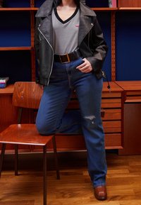 Černá kožená motorkářská bunda, proužkované tričko s výstřihem do V, modré džíny s vysokým pasem a dírou, hnědé kotníkové boty na blokovém podpatku. Dřevěné pozadí.