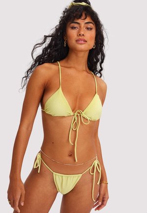 Femme aux cheveux longs et bouclés portant un bikini jaune clair à fines bretelles nouées, des boucles d'oreilles anneaux dorés, et une fine chaîne de taille sur un fond uni.