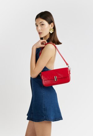 Giovane donna con vestito di jeans blu senza maniche che porta una borsa a tracolla rossa in pelle e indossa grandi orecchini floreali dorati.