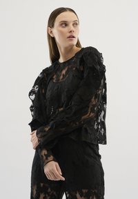 Top din dantelă neagră cu un model floral texturat, mâneci volan pentru un efect elegant și o suprapunere transparentă. Mâneci lungi cu manșete ajustate și decolteu rotund.
