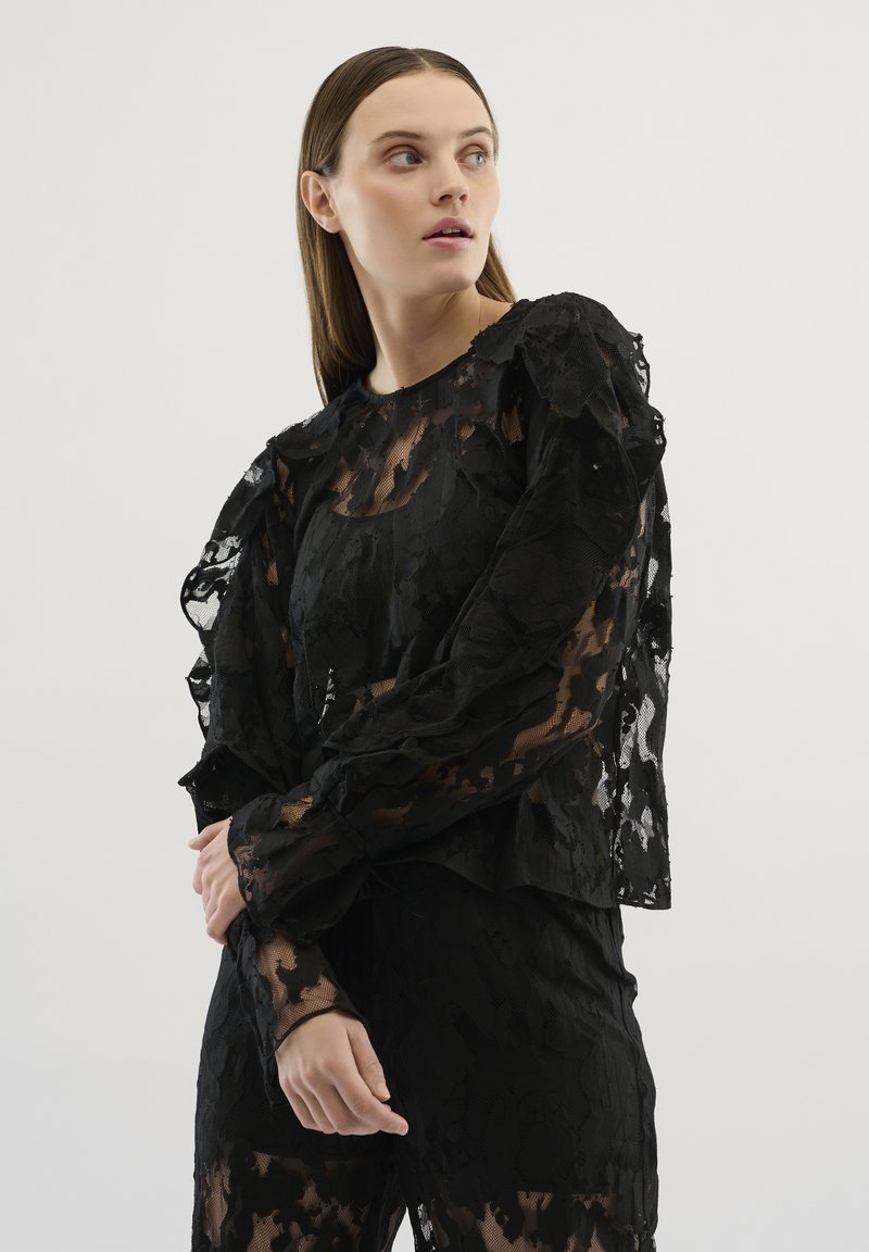Top din dantelă neagră cu un model floral texturat, mâneci volan pentru un efect elegant și o suprapunere transparentă. Mâneci lungi cu manșete ajustate și decolteu rotund.