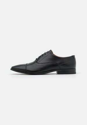 Chaussure Oxford pour homme en cuir noir avec laçage fermé, bout pointu et talon bas sur un fond blanc uni.