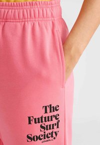 Rosa bomullsbyxor med resårmidja, sidofickor och fet svart text som läser "The Future Surf Society" på låret.