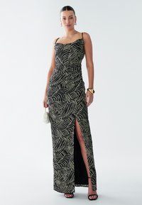 Vestido negro con un patrón de olas en dorado, con tirantes finos, una abertura lateral y un diseño ajustado. La modelo sostiene un pequeño bolso claro.