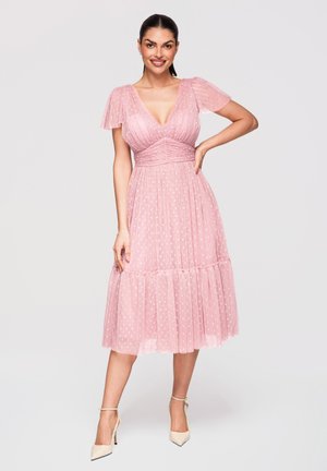 Femme souriante portant une robe midi rose à pois avec des manches volantées et des talons beige à bride à la cheville, debout devant un fond uni.