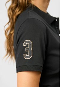 Zwarte poloshirt van textiel met een gestructureerde stof, voorzien van korte mouwen en een geborduurd nummer "3" in grijs en beige op de bovenarm.