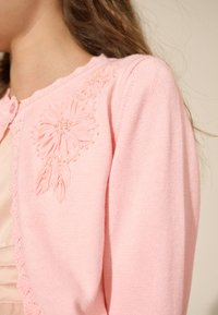 Cardigan rosa chiaro con ricamo floreale e dettagli di perline sulla spalla sinistra, con bordo smerlato lungo la chiusura con bottoni.