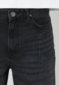 Jean en denim noir avec une finition texturée, présentant une fermeture à bouton sur le devant, cinq poches et des passants de ceinture.