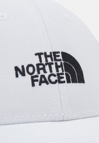 Balta skrybėlė, pagaminta iš audinio, su juodu siuvinėtu logotipu "THE NORTH FACE" ir puslanka dizainu. Siūlės matomos aplink siūles.