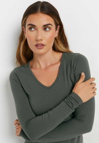 Maglia lunga verde scuro con design a V. Realizzata in un tessuto liscio e leggero. Maniche aderenti con leggera arricciatura ai polsi.