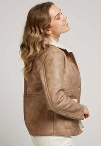 Veste en cuir synthétique marron avec un col en fourrure synthétique crème, poches latérales et détails de couture visibles ; texture lisse et design structuré.
