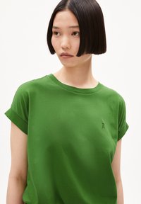 Camiseta de algodón verde con mangas cortas enrolladas. Presenta un pequeño logotipo bordado en el pecho. Textura suave, cuello redondo y corte relajado.