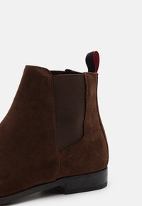 Bottine en daim marron avec panneaux latéraux élastiques, un passant de talon en tissu contrastant et une semelle en caoutchouc noire. Design simple et minimaliste.