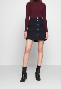 Jersey de cuello alto burdeos con canalé combinado con una minifalda negra de línea A con bolsillos delanteros y botones plateados. Botines negros de tacón.