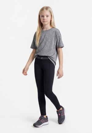 Ung jente står iført en grå t-skjorte, svarte leggings og grå og rosa treningssko mot en hvit bakgrunn.