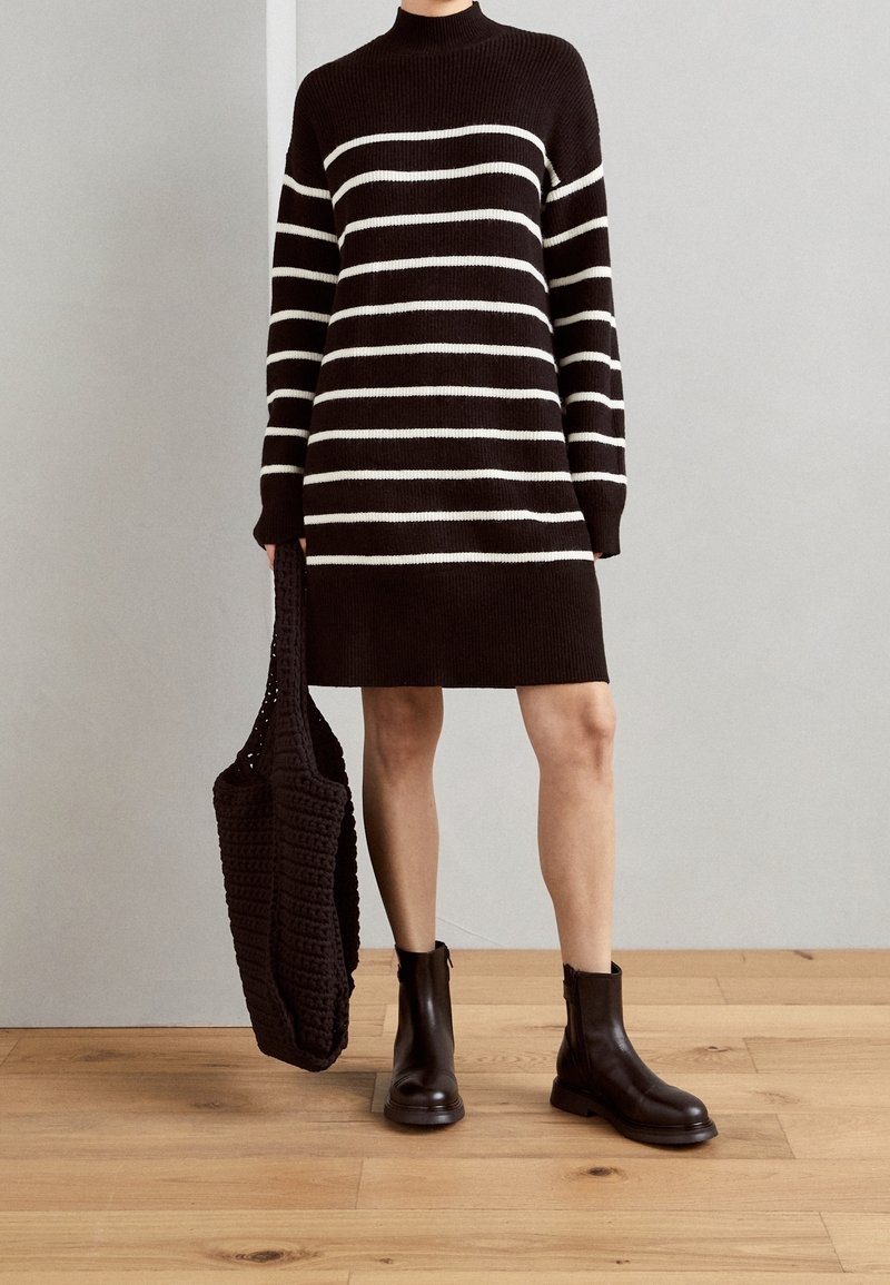 Personne portant une robe pull à rayures horizontales noires et blanches, des bottines noires, et tenant un sac fourre-tout en maille noire.