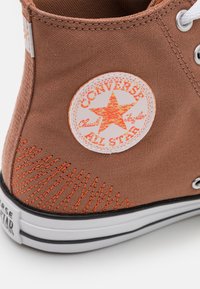 Sapatilha alta de lona castanha com um destaque bordado laranja e um logótipo circular branco com uma estrela, sola de borracha preta.