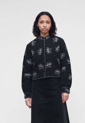 Femme aux cheveux bruns mi-longs porte une veste noire texturée avec des motifs floraux argentés et une jupe noire sur un fond uni.