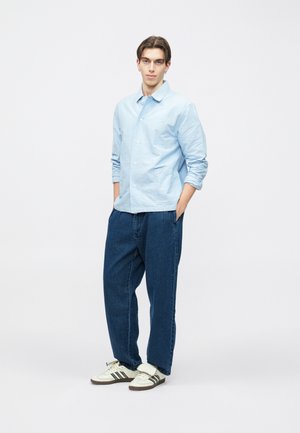 Camicia celeste chiusa con bottoni, pantaloni di jeans blu dalla vestibilità comoda e sneakers bianche con strisce nere. Il modello è in piedi con le mani nelle tasche, sfondo neutro.