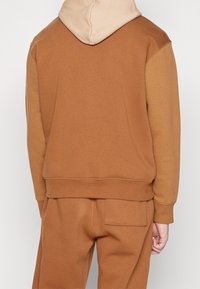 Personne portant un sweat à capuche bicolore marron et un pantalon marron assorti, vue de dos avec une main légèrement visible.