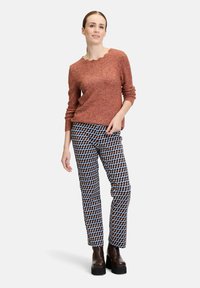 Rotbrauner Pullover mit gewelltem Ausschnitt, kombiniert mit gemusterten Hosen in Blau, Braun und Weiß. Klobige, braune Stiefeletten mit Absatz.