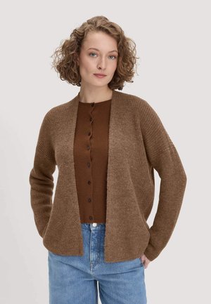 Frau mit kurzen lockigen Haaren, die eine braune Strickjacke über einem braunen, geknöpften Top und blauen Jeans trägt, steht mit den Händen in den Taschen.